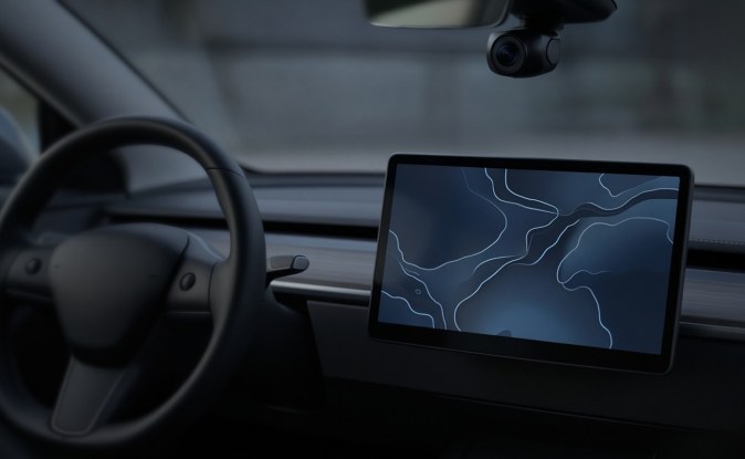Interior de un auto eléctrico con volante, pantalla central con un mapa abstracto y una cámara bajo el espejo retrovisor.