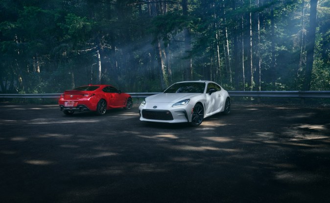 Dos Toyota GR86, uno rojo y uno blanco, estacionados en un camino rodeado de bosque.
