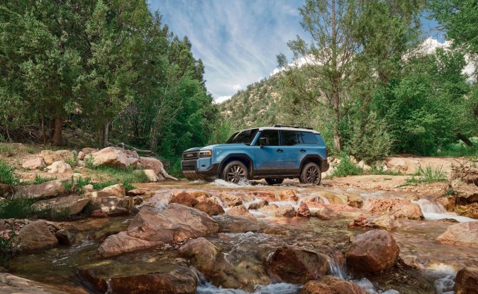 Toyota Land Cruiser azul cruzando un arroyo rocoso entre árboles, vista de lado.
