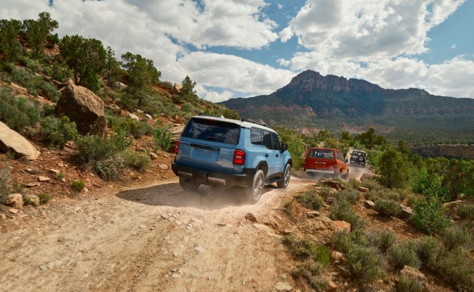 Toyota Land Cruiser azul subiendo un camino de tierra, vista desde atrás, con montañas al fondo.