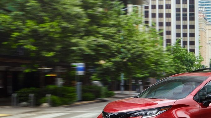 Minivan Toyota Sienna roja de frente en una calle urbana, con edificios y árboles al fondo.
