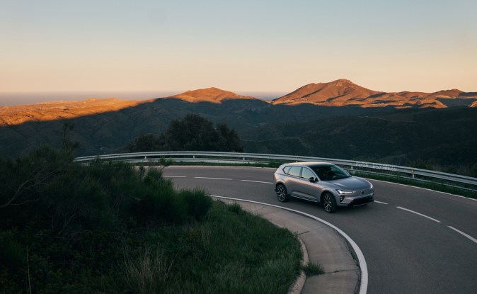 Volvo EX60 plateado circulando por una carretera de montaña al atardecer con un valle al fondo
