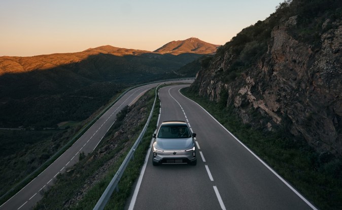 Vista elevada del Volvo EX60 plateado bajando por una curva en un puerto de montaña al atardecer