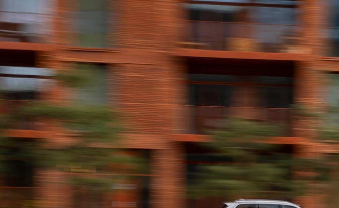 SUV Volvo XC90 plateado circulando junto a un canal frente a edificios de ladrillo.